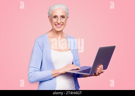 Grey haired old nice beautiful cheerful smiling woman wearing spectacles holding laptop learning online course. Isolated over violet purple background Stock Photo