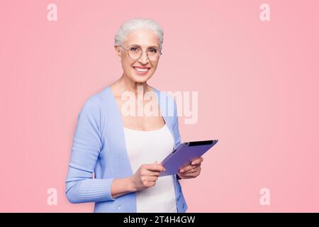 Grey haired old smiling business woman wearing glasses, reading browsing using holding tablet. Isolated over violet purple background Stock Photo