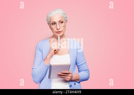 Gray haired old serious business woman wearing glasses, thinking about plans strategy future, holding paper note book with pen. Isolated over violet Stock Photo