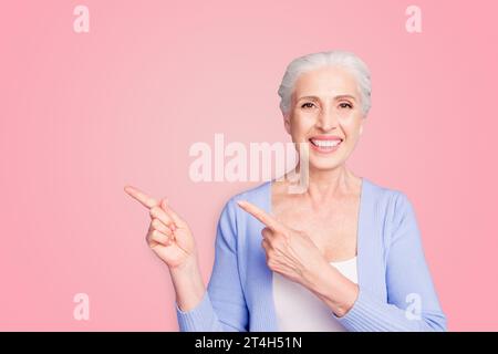 Beautiful nice cheerful smiling gray-haired old lady wearing casual, showing pointing way with two fingers. Copy space. Isolated over pastel violet Stock Photo