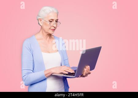 Gray haired old serious business woman wearing glasses, holding computer, typing texts, browsing. Isolated over violet purple background Stock Photo