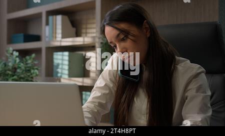 Caucasian businesswoman talking phone typing laptop multitask in office business girl woman using computer answering call talk smartphone consult Stock Photo