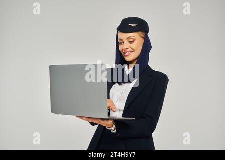 pleased arabian airlines stewardess in corporate uniform using laptop while standing on grey Stock Photo