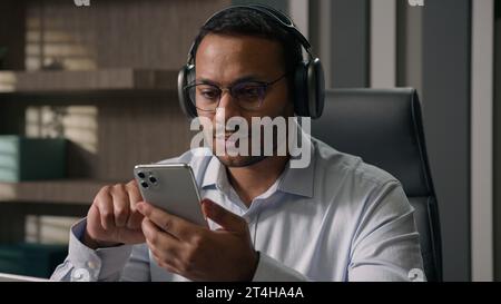 American businessman Indian man Arabian worker boss in headphones listen music using mobile phone choose song in sound app at office table calm audio Stock Photo
