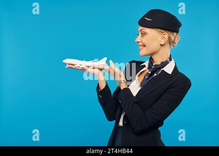 cheerful attractive stewardess in uniform standing with airplane model on blue backdrop, side view Stock Photo