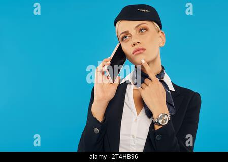 thoughtful air hostess in uniform talking on mobile phone and looking away on blue background Stock Photo