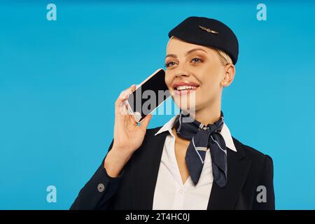 young and cheerful stewardess in elegant uniform talking on smartphone with blank screen on blue Stock Photo
