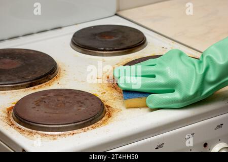 Woman hand in glove remaining burnt stains on white dirty electric stove scrubbing using sponge Stock Photo