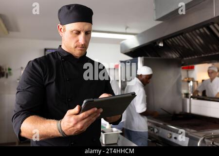Diverse male chef teacher working over tablet while student cooking food in commercial kitchen Stock Photo