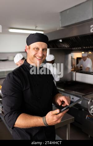 Diverse smiling chef teacher in black uniform using tablet while students working in background Stock Photo