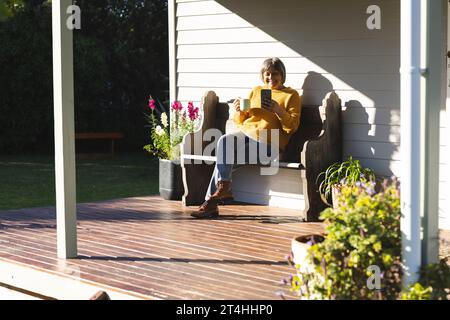 Happy senior biracial woman drinking tea and using smartphone sitting in garden, copy space Stock Photo