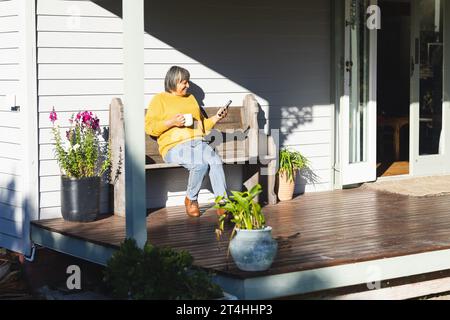 Happy senior biracial woman drinking tea and using smartphone sitting in garden, copy space Stock Photo