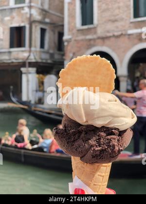 ice cream, closeup side view female hand holds delicious ice cream scoops in waffle cone over blurred people in iconic gondola on canal of Venezia or Stock Photo