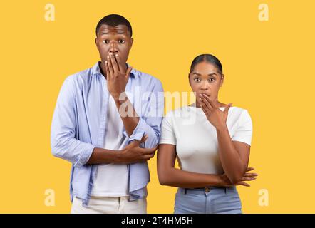 Shocking news. Young black couple man and woman with wide open eyes, covering mouth with hand, yellow background Stock Photo