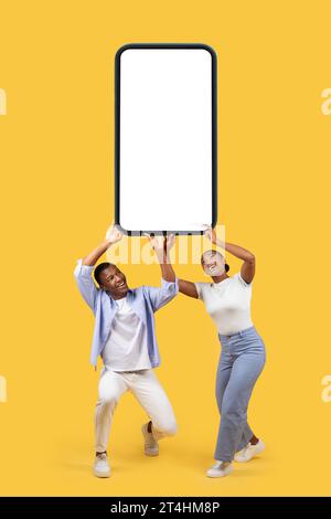 Black couple holding huge heavy smartphone with white black screen on yellow studio background, mockup Stock Photo