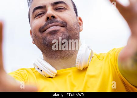 A cheerful man, wearing a vibrant t-shirt and wireless headphones, playfully poses with a taking selfie on camera Stock Photo