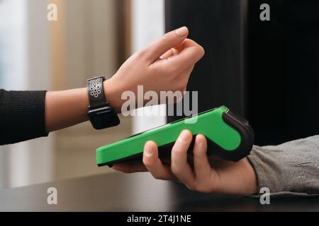 Closeup of modern lady paying via smartwatch while making purchase in shop. Woman customer using hand watch for contactless payment via payment terminal on counter in store. NFC technology concept Stock Photo