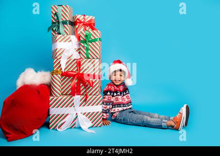Photo of satisfied small boy with curly hair dressed ornament sweater santa hat sit near surprises isolated on blue color background Stock Photo