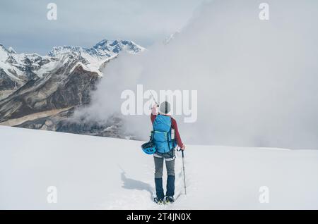 Climber with backpack and trekking poles descending Mera peak high slopes at cca 6000m and pointing at legendary Mount Everest, Nuptse, Lhotse with So Stock Photo