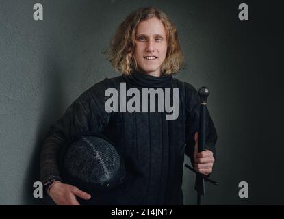 Portrait of a smiling long-haired young teen dressed in black historical fencing armor with a long medieval sharp sword Stock Photo