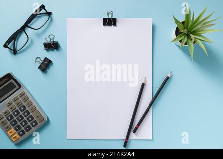 Top view of blank notebook, glasses, pen, calculator and green plant on blue office desk Stock Photo