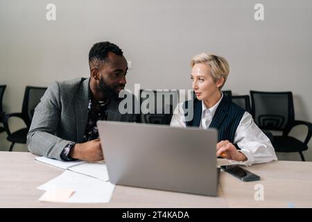 Positive African male and Caucasian female business employees cooperating in office, smiling talking working together at workplace in office. Stock Photo