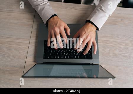 Close-up top view of unrecognizable businessman in suit working on laptop computer by window. Closeup male fingers typing buttons on laptop keyboard. Stock Photo