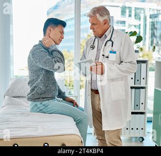 Mature medical doctor standing and using a digital tablet while checking on his injured asian patient in a clinic. Young mixed race man suffering from Stock Photo