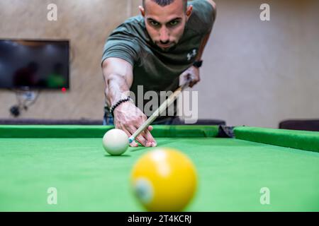 Young adult bearder male playing billiards with his friends focusing on white ball to hit it with muscular hand Stock Photo