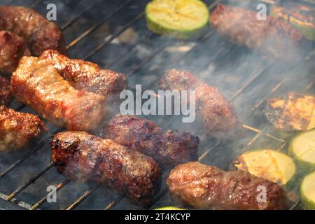 Grilling meat rolls called mici or mititei with vegetables on char barbecue. Charcoal grill with burning fire Stock Photo