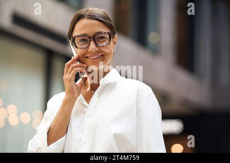 Cheerful woman entrepreneur talking phone with client while standing on office background Stock Photo