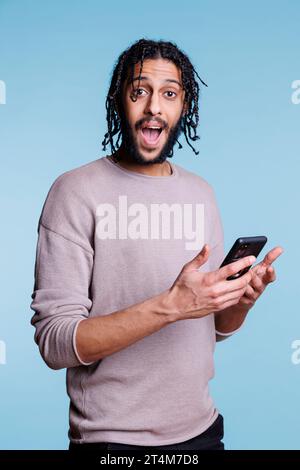 Excited arab man using smartphone and looking at camera with happy facial expression. Cheerful person in casual clothes getting online message with good news on mobile phone portrait Stock Photo