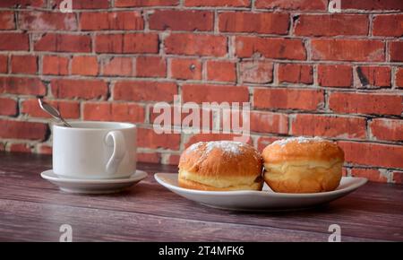 A cup of hot coffee on a saucer and a plate with two fresh donuts topped with powdered sugar on a wooden counter against a brick wall. Close-up. Stock Photo