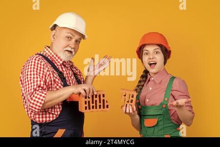 amazed child and grandfather bricklayer in helmet on yellow background ...