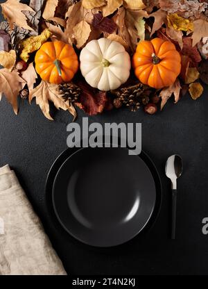 Pumpkin soup preparation concept. Festive autumn table setting with miniature pumpkins over autumn leaves and an empty black soup bowl or plate over b Stock Photo