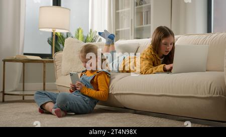 Children and modern technology two happy kids girls sisters friends play video online game together on laptop phone in cozy living room having fun Stock Photo