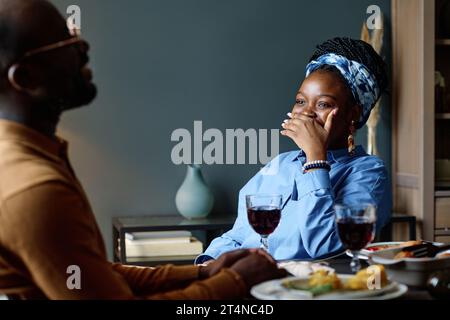Young laughing woman covering her mouth by hand and looking at her husband sitting in front of her by festive table with glasses of wine Stock Photo