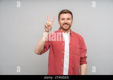 Smiling positive man showing victory sign looking at camera on gray background. Stock Photo