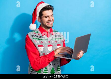Photo portrait of cute young man working laptop worker freelance wear x-mas print vest hat clothes isolated on blue color background Stock Photo