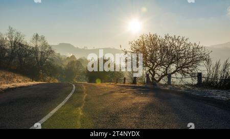 road through mountainous countryside in morning light. leafless tree on the hills. sun on the blue cloudless sky Stock Photo