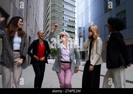 Happy business senior woman dancing outside office building with female colleagues celebrating. Stock Photo
