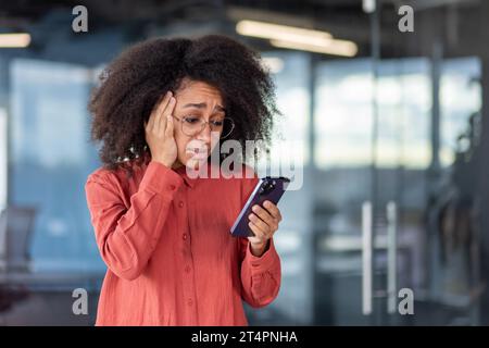 Businesswoman received notification on phone about bad news, bankruptcy and loss of money, woman at workplace inside office, using app on smartphone, disappointed and sad. Stock Photo