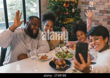 Cheerful african american family enjoying christmas and new years lunch while woman taking selfie on smartphone with members of family at home against decorated xmas tree Stock Photo