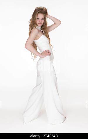 Woman in white long jumpsuit with one hand behind head and other hand on waist standing white background. Young adult fashion model with long hair Stock Photo