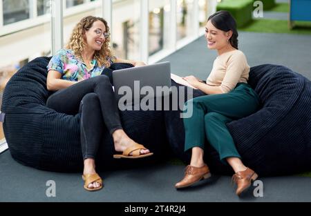 Maintaining innovation through their collaboration. two businesswomen using a laptop together while sitting on beanbags in an office. Stock Photo