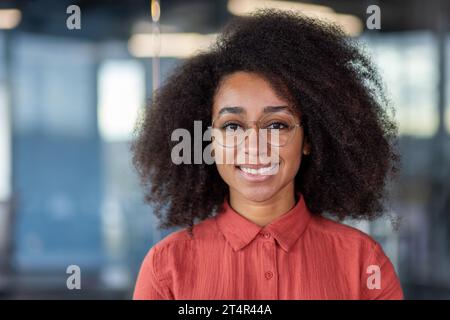 Close-up portrait of woman inside office, businesswoman happy and smiling looking at camera, female worker in red shirt and curly hair, satisfied Latin American programmer developer. Stock Photo