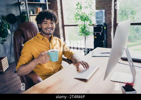 Photo portrait of attractive young man wear yellow shirt working manager sit table hold coffee cup modern workplace room home design Stock Photo