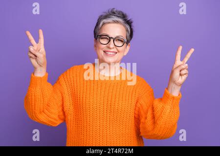 Portrait photo of senior businesswoman wearing orange sweater showing double v sign friendly greetings isolated on violet color background Stock Photo