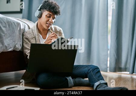 Stay at home, slay your goals. a young man writing in a notebook and using a laptop while listening to music at home. Stock Photo
