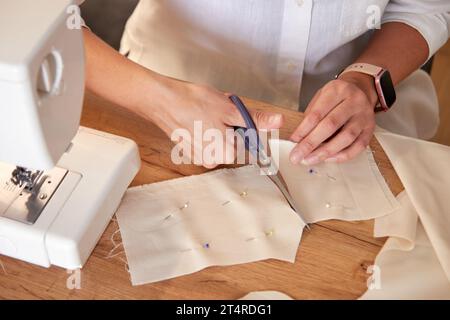 Hands of seamstress using scissors cutting beige fabric with pinned small cutouts. Creative young adult woman making handmade dolls. Stock Photo